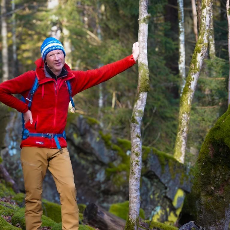 Ein Wanderer in roter Jacke steht in einem Wald und zeigt mit der Hand auf einen bemoosten Baumstamm.